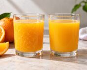 Dos vasos de zumo de naranja, uno con pulpa visible y otro de textura uniforme, junto a naranjas